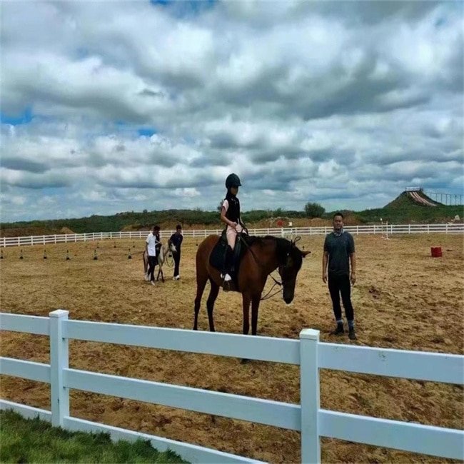 A white PVC fence with three horizontal rails surrounding a horse riding arena. A young rider on horseback is accompanied by an instructor, while other riders and horses are visible in the background. The arena is set against a backdrop of a partly cloudy sky and open landscape.