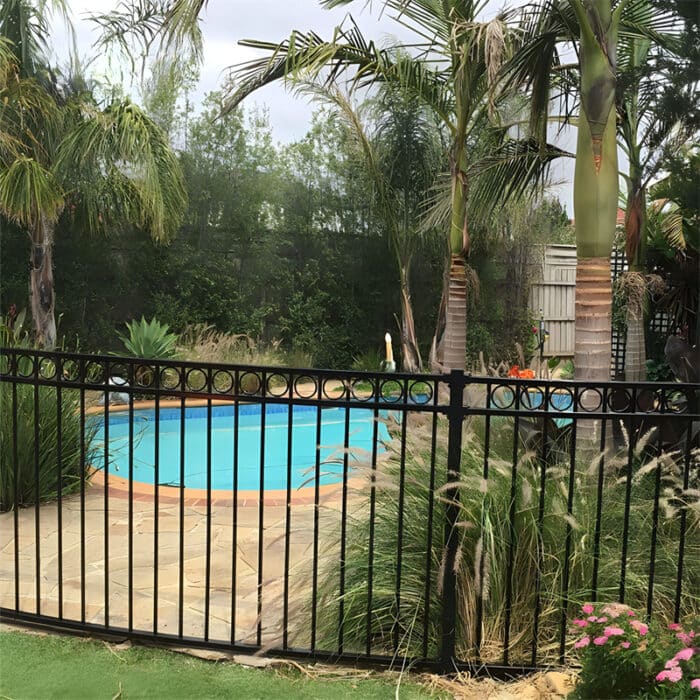 Black aluminum flat top fence with decorative rings installed around a backyard swimming pool, surrounded by lush greenery and palm trees, providing safety and a stylish appearance.