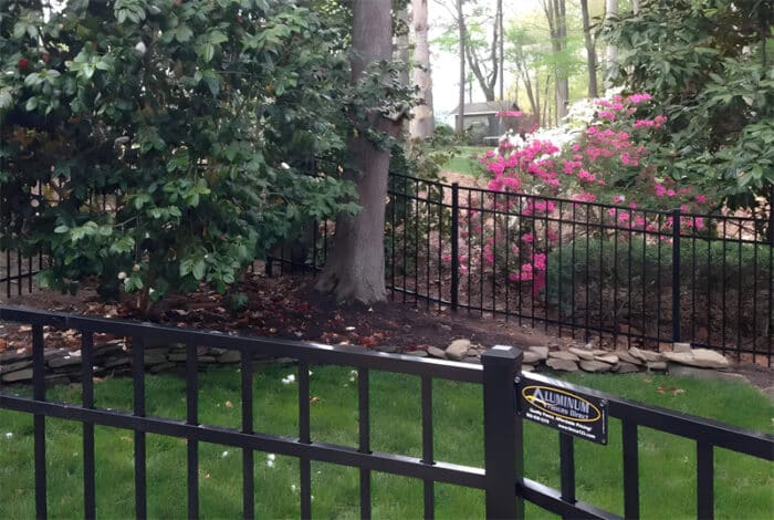 Black aluminum fence installed around a lush garden with blooming flowers and tall trees, enhancing both security and aesthetics.