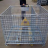 Worker maneuvering a pallet jack with a collapsible stillage cage in an outdoor storage area.