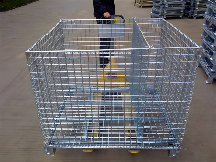 Worker maneuvering a pallet jack with a collapsible stillage cage in an outdoor storage area.