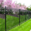 A sleek black aluminum fence panel with vertical bars and decorative top accents, installed along a lush green lawn with blooming pink trees in the background.