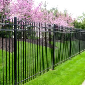 A sleek black aluminum fence panel with vertical bars and decorative top accents, installed along a lush green lawn with blooming pink trees in the background.
