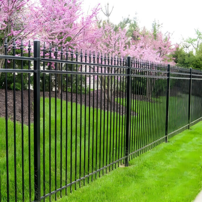 Un elegante panel de cerca de aluminio negro con barras verticales y detalles decorativos en la parte superior, instalado a lo largo de un exuberante césped verde con árboles rosados en flor en el fondo.