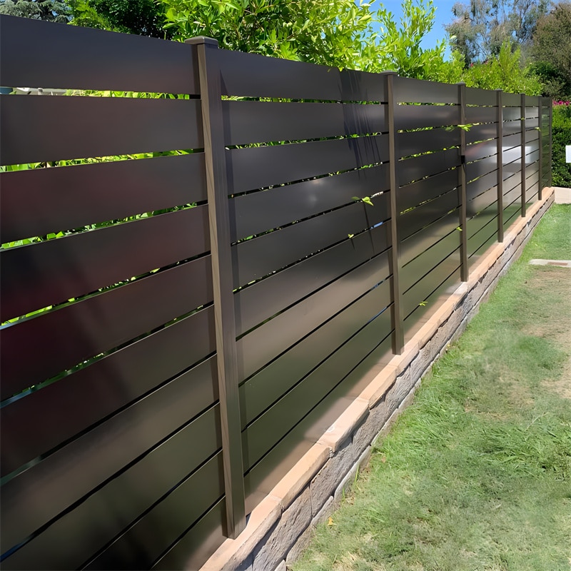 Modern black aluminium slat fence installed in a garden, providing a sleek and stylish privacy solution. The fence features horizontal slats with a smooth finish, supported by vertical posts and set on a stone base.