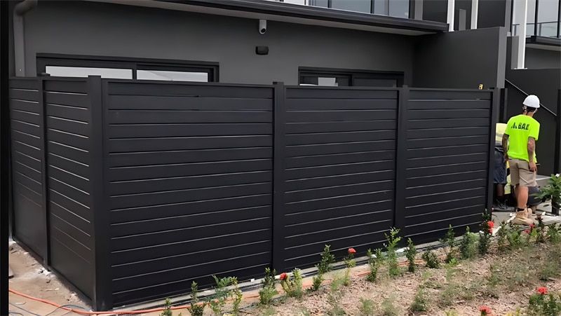 Elegant black aluminium slat fence installed in a garden, featuring horizontal slats and vertical posts on a stone base.