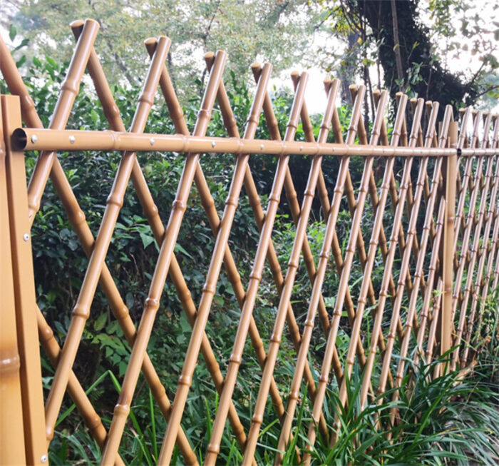 An outdoor installation of tan-colored stainless steel bamboo-style fencing with diamond latticework, blending seamlessly into a lush, green landscape.