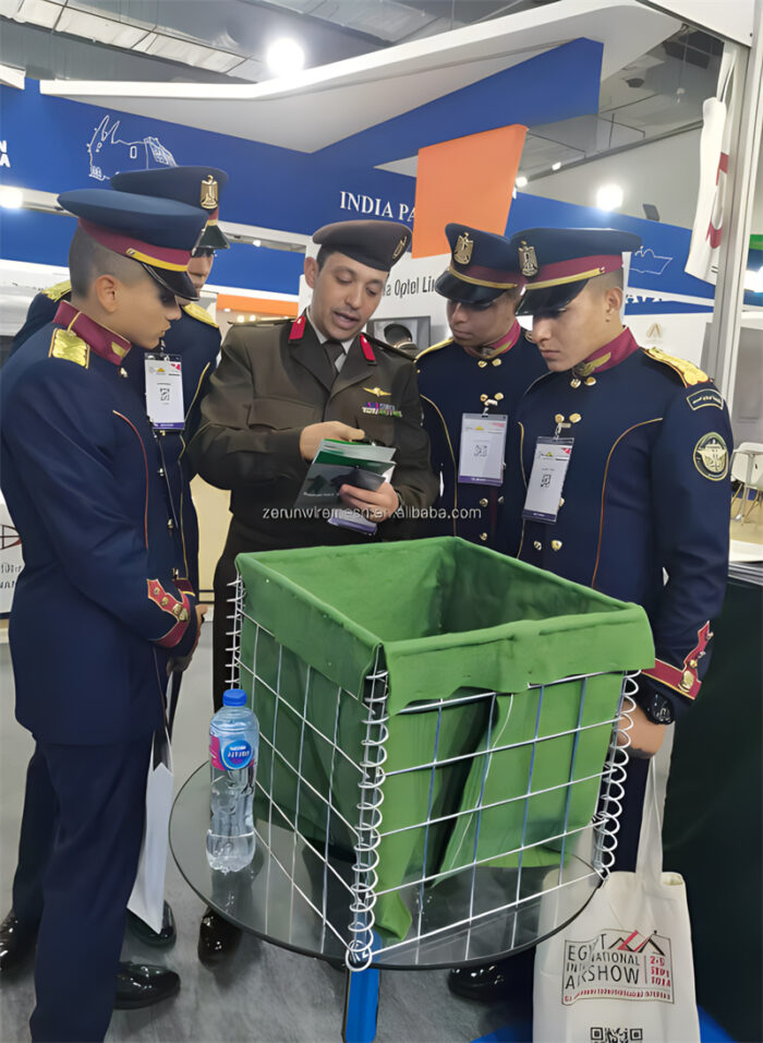 Military personnel discussing the assembly of a green Hesco barrier.