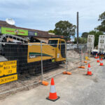 Construction machinery behind temporary fencing, with warning signs for pedestrians in the foreground.