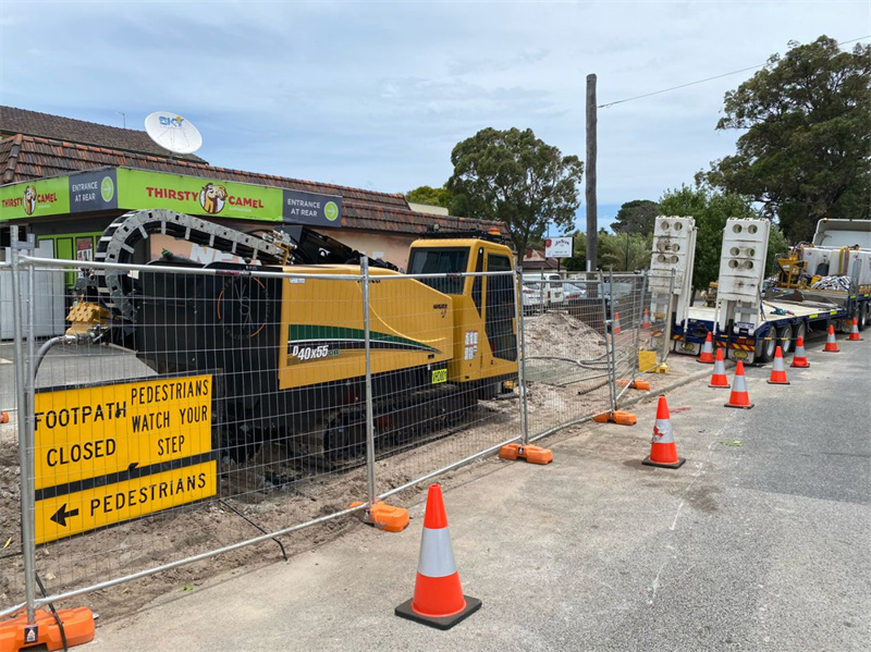 Construction machinery behind temporary fencing, with warning signs for pedestrians in the foreground.