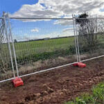 Temporary fencing set up on a rural site, with red concrete feet holding the panels in place against a green backdrop.