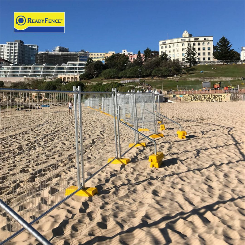 Temporary fencing on the beach with yellow bases, near buildings and grassy area.