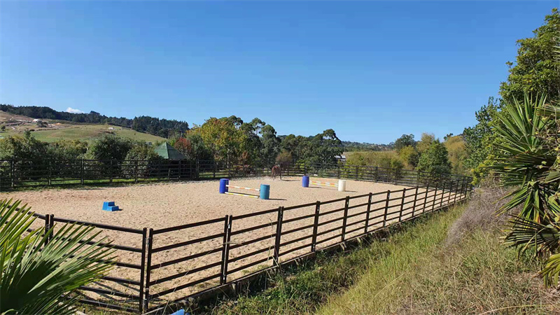 A well-maintained horse training yard featuring sand and obstacles in a rural setting with trees in the background.