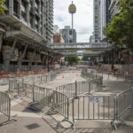 A view of a city street with a complex arrangement of steel crowd control barriers, directing pedestrian traffic and maintaining safety during construction activities, with a tower visible in the background.