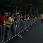 A group of people standing by silver barricades watching a marathon, indicating a lively event atmosphere.