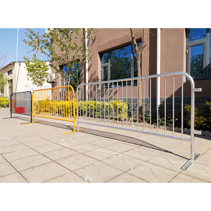 A row of steel crowd barriers, including a yellow barrier, placed on a paved walkway surrounded by greenery, providing crowd control and safety.