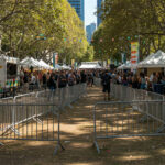 A busy outdoor festival scene showing rows of crowd control barricades directing foot traffic along a pathway lined with food stalls and tents.