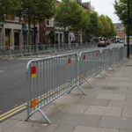 A row of steel barricades set along a city street, creating a safe pedestrian area and preventing vehicle access, with shops and trees in the background.