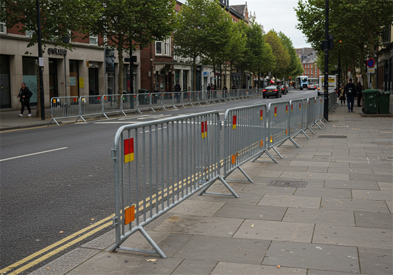 A row of steel barricades set along a city street, creating a safe pedestrian area and preventing vehicle access, with shops and trees in the background.