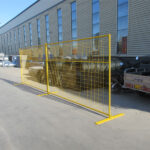 Yellow temporary fencing panels with flat feet, set up outdoors in front of an industrial building.