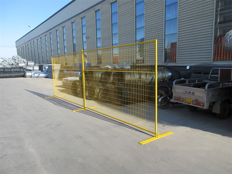 Yellow temporary fencing panels with flat feet, set up outdoors in front of an industrial building.