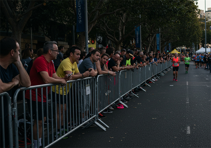 A group of people standing by silver barricades watching a marathon, indicating a lively event atmosphere.