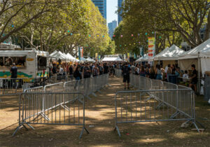 A busy outdoor festival scene showing rows of crowd control barricades directing foot traffic along a pathway lined with food stalls and tents.