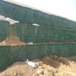 A metal mesh barrier wall covered in colorful graffiti with tags like "MAKS" and "THE COLORADO KILLER" in red, blue, white, and green spray paint. The wall is set in an urban construction site with gravel and debris on the ground.