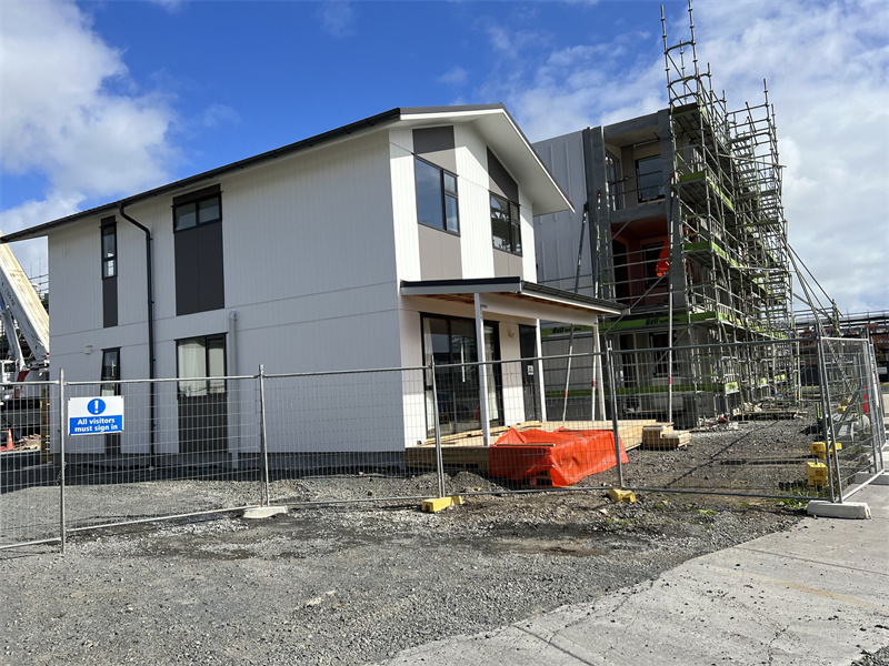 A newly constructed two-story house is surrounded by a temporary fence, with a partially constructed building behind it covered in scaffolding. A sign on the fence reads "All visitors must report."