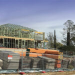 A building under construction with exposed framing, surrounded by fencing and construction materials stored on-site.