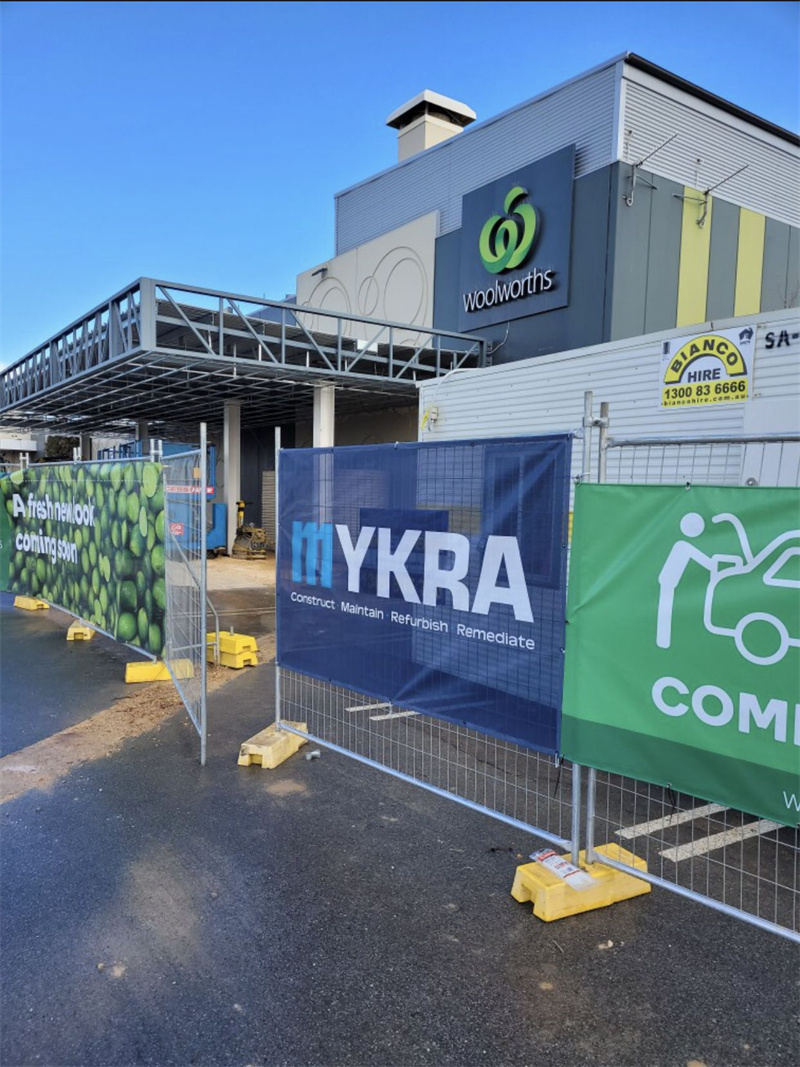 A construction site fenced off with banners displaying branding and safety messages, located outside a Woolworths store, with construction equipment visible in the background.