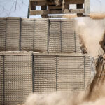 A close-up of military defensive barriers, filled with sand and reinforced with wire mesh, as machinery operates nearby, creating dust in the air.