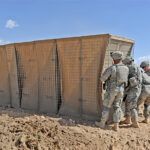 A group of soldiers installing a Hesco barrier in a desert environment for protective reinforcement and perimeter security.