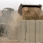 A military vehicle unloading a large pile of sand into a metal mesh barrier in a desert environment, creating a protective wall. Dust is rising from the sand as it pours, and the landscape is barren with scattered debris in the background.