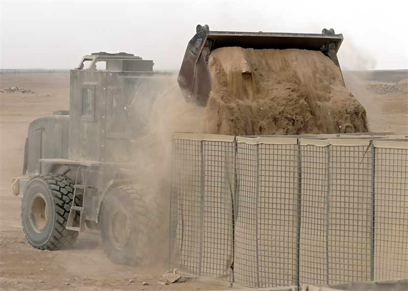 A military vehicle unloading a large pile of sand into a metal mesh barrier in a desert environment, creating a protective wall. Dust is rising from the sand as it pours, and the landscape is barren with scattered debris in the background.