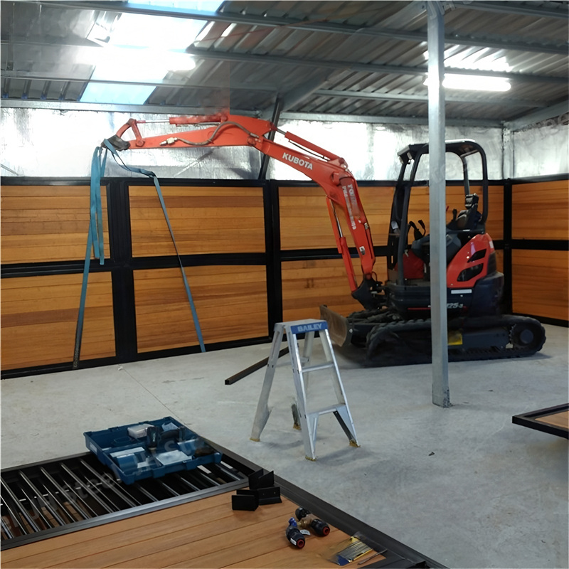 A construction site for horse stalls, featuring a small excavator and tools, with wooden panels being installed in the background.