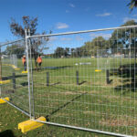 A construction site with temporary fencing around a grassy area. Workers in orange vests are visible in the background, and the scene is set against a clear blue sky with a few clouds.