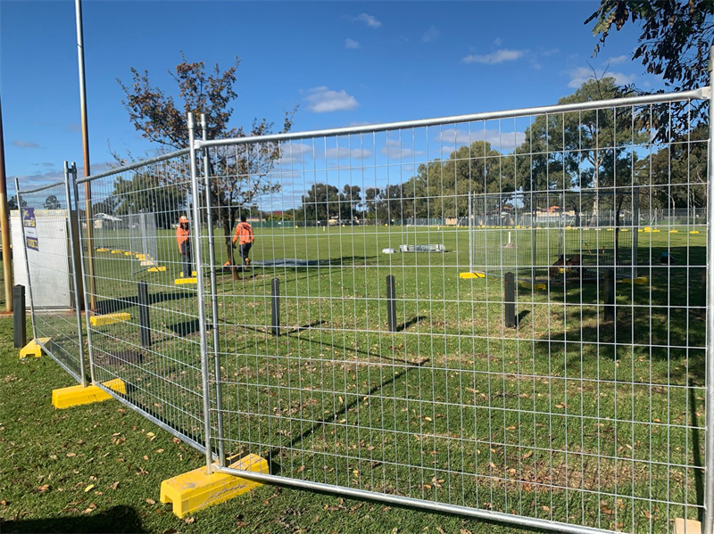 A construction site with temporary fencing around a grassy area. Workers in orange vests are visible in the background, and the scene is set against a clear blue sky with a few clouds.