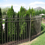 An elegant black wrought iron fence with gold finials, surrounding a luxurious building with stone pillars and green landscaping, showcasing a grand entrance.