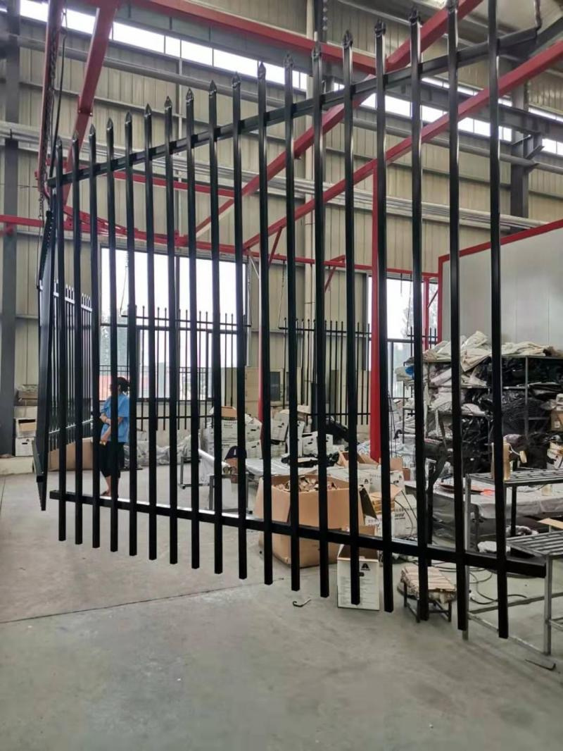 A partially assembled black metal fence in a workshop environment, with boxes and materials scattered around. A worker is seen in the background.