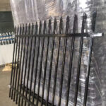 A black metal fence with pointed tops leaning against a stack of wrapped fence panels. The warehouse interior is visible in the background, indicating preparation for shipping or installation.
