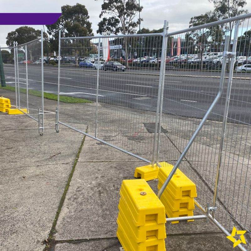 A fenced area with yellow barriers, showing a construction site with vehicles and materials in the background.