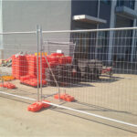 A construction site fenced with a metal barrier, featuring orange base supports, with construction materials stacked nearby.