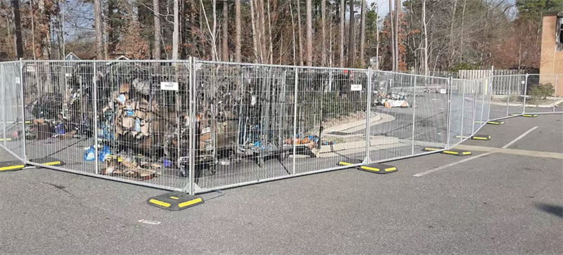 A fenced-off area containing debris and equipment, enclosed by chain-link fencing supported by multiple yellow-tipped gray plastic bases, situated in a parking lot surrounded by trees.