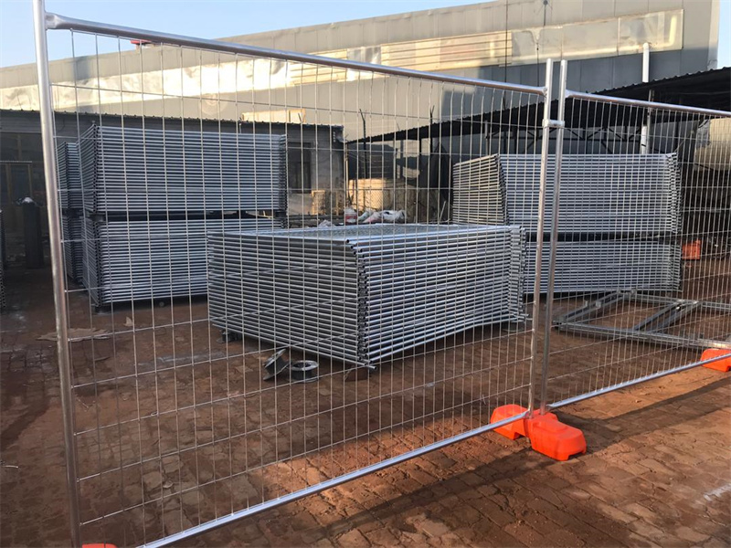 A fenced-off area containing stacked metal panels, secured by orange bases. The background shows a construction site with various materials scattered around, indicating ongoing work.