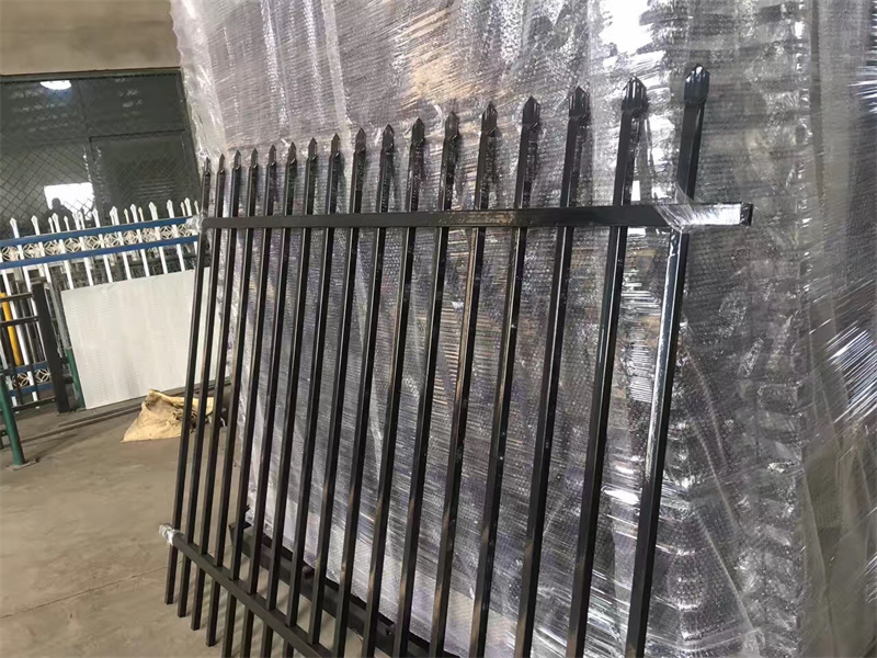A black metal fence with pointed tops leaning against a stack of wrapped fence panels. The warehouse interior is visible in the background, indicating preparation for shipping or installation.