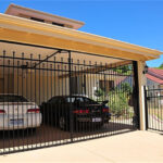 A black metal gate is shown in front of a carport where two cars are parked. The gate features decorative spikes and is positioned under an awning, with a clear blue sky in the background.