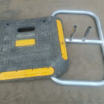 A close-up of a heavy-duty plastic base with yellow edges next to an aluminum frame, which is used to support fencing, lying on a sandy surface.
