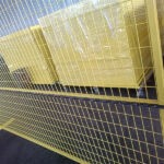 A bright yellow wire mesh fence stands in a warehouse, with neatly stacked yellow panels visible in the background and a tape measure being held in front to show the fence's dimensions.