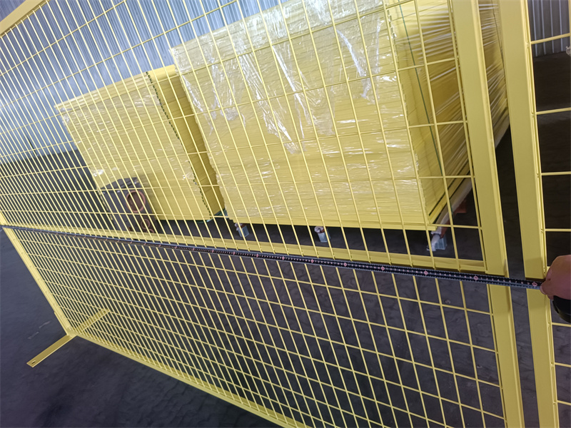 A bright yellow wire mesh fence stands in a warehouse, with neatly stacked yellow panels visible in the background and a tape measure being held in front to show the fence's dimensions.